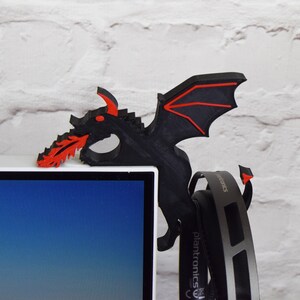 May include: Black and red dragon-shaped headset holder. The dragon is perched on the corner of a computer monitor, with its wings and fiery breath extending outwards. The headset hangs below the dragon.