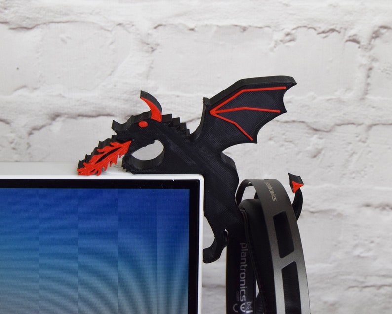 May include: Black and red dragon-shaped headset holder. The dragon is perched on the corner of a computer monitor, with its wings and fiery breath extending outwards. The headset hangs below the dragon.
