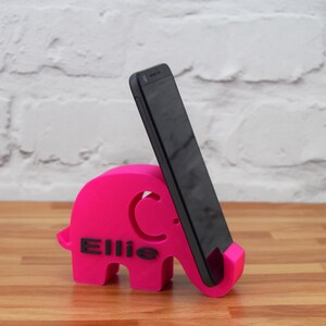 Pode incluir: Um suporte de telefone de elefante rosa com o nome "Ellie" impresso nele. Um smartphone preto est&aacute; em p&eacute; no suporte.