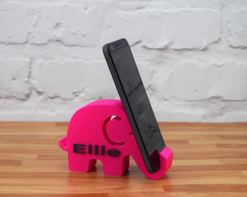 Pode incluir: Um suporte de telefone de elefante rosa com o nome "Ellie" impresso nele. Um smartphone preto est&aacute; em p&eacute; no suporte.