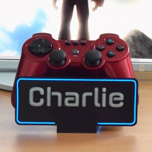 Pu&ograve; includere: Un controller per videogiochi rosso con una base nera e un cartello al neon blu che dice "Charlie".