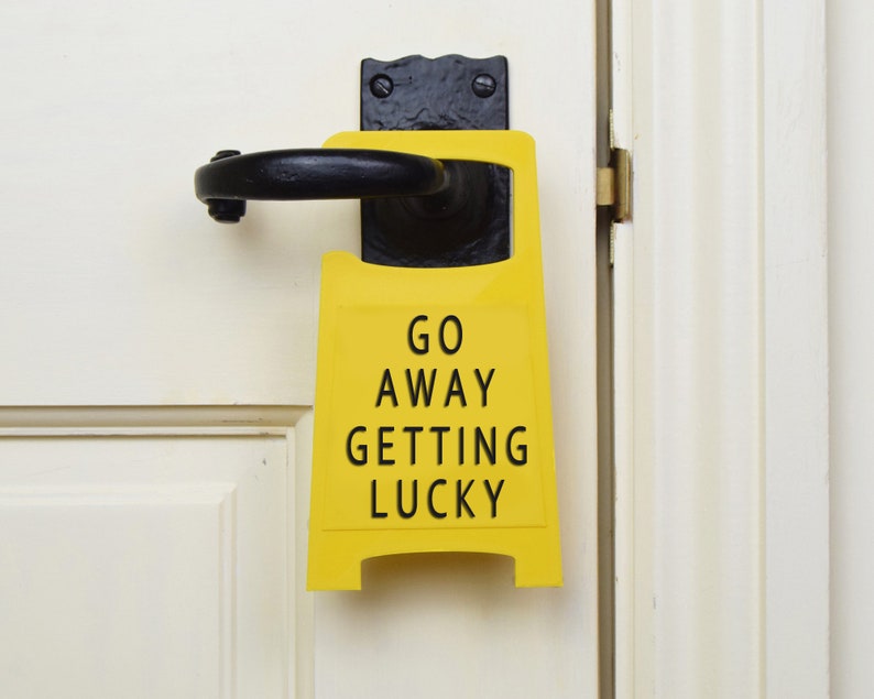 Op de afbeelding: Een geel waarschuwingsbord met zwarte tekst dat "Go Away Getting Lucky" zegt, hangend aan een witte deur.