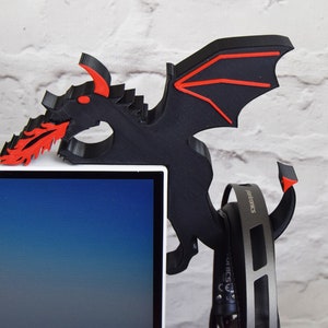 May include: A black and red dragon-shaped headphone holder. The dragon is mounted on a white computer monitor. The dragon has red accents on its wings and mouth, which is spewing red flames. A pair of headphones hangs from the holder.