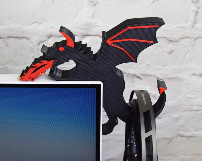 May include: A black and red dragon-shaped headphone holder. The dragon is mounted on a white computer monitor. The dragon has red accents on its wings and mouth, which is spewing red flames. A pair of headphones hangs from the holder.