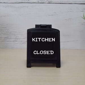 May include: A black chalkboard sign displaying the words "KITCHEN CLOSED" in white. The sign is in an A-frame design and rests on a light wooden surface. A decorative plant in a pot is visible in the background.