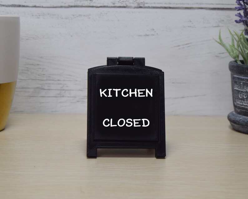May include: A black chalkboard sign displaying the words "KITCHEN CLOSED" in white. The sign is in an A-frame design and rests on a light wooden surface. A decorative plant in a pot is visible in the background.