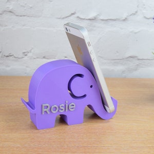 Pode incluir: Suporte de telefone de elefante roxo com o nome "Rosie" e a letra "C" no lado. O suporte est&aacute; segurando um smartphone prateado.