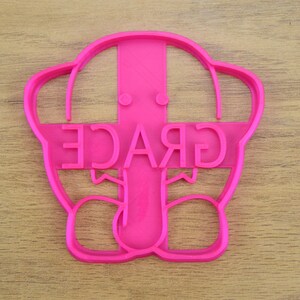 May include: Pink plastic cookie cutter shaped like a cartoon elephant with the word "GRACE" written across the elephant's head.