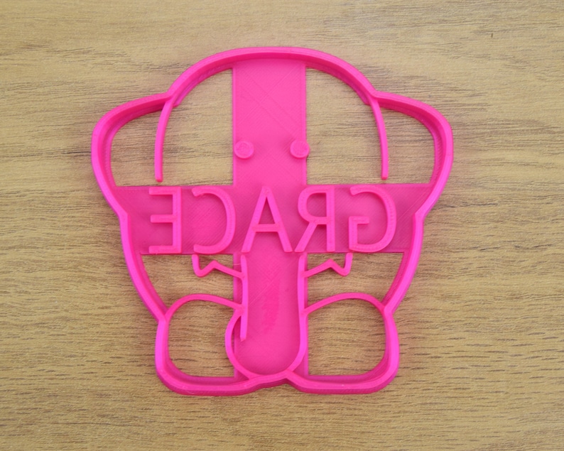 May include: Pink plastic cookie cutter shaped like a cartoon elephant with the word "GRACE" written across the elephant's head.