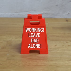 May include: Red plastic sign with the text "WORKING! LEAVE DAD ALONE!" on a wooden surface.