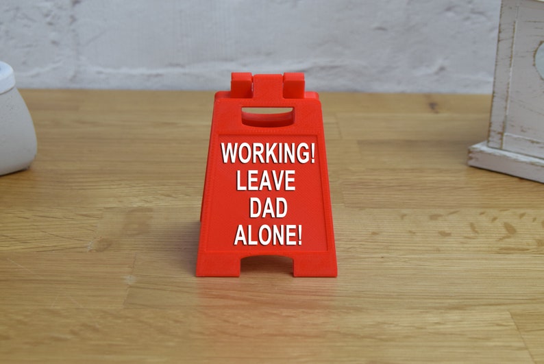 May include: Red plastic sign with the text "WORKING! LEAVE DAD ALONE!" on a wooden surface.