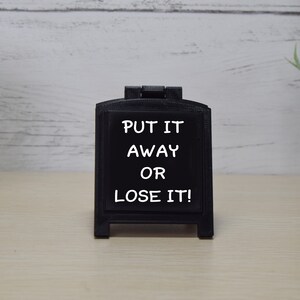 May include: A small, black chalkboard sign with the text "PUT IT AWAY OR LOSE IT!" in white. The sign is in a small, decorative frame. The background is a light-coloured wooden surface.