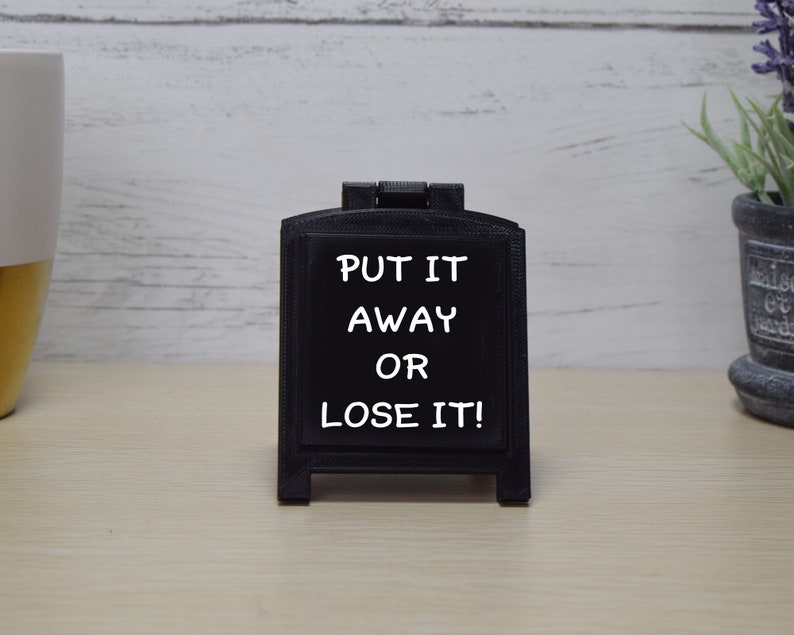 May include: A small, black chalkboard sign with the text "PUT IT AWAY OR LOSE IT!" in white. The sign is in a small, decorative frame. The background is a light-coloured wooden surface.