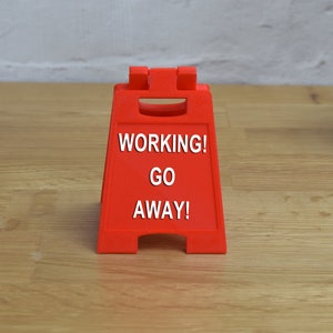 May include: A red plastic sign with the text "WORKING! GO AWAY!" on a wooden surface.