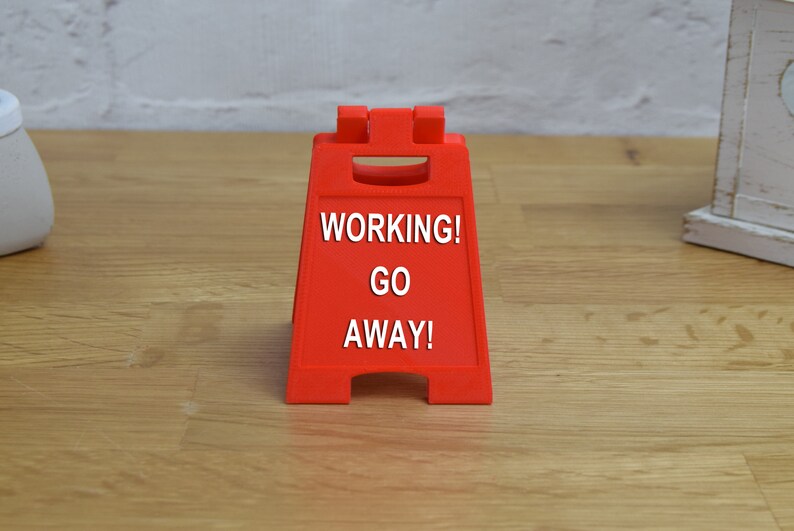 May include: A red plastic sign with the text "WORKING! GO AWAY!" on a wooden surface.