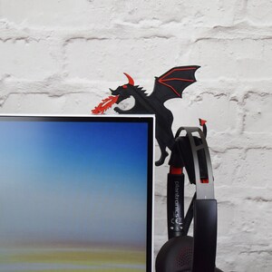 May include: A black dragon-shaped desk accessory with red accents, perched on the edge of a computer monitor. The dragon is spewing red flames. A black and silver headset hangs below the dragon.