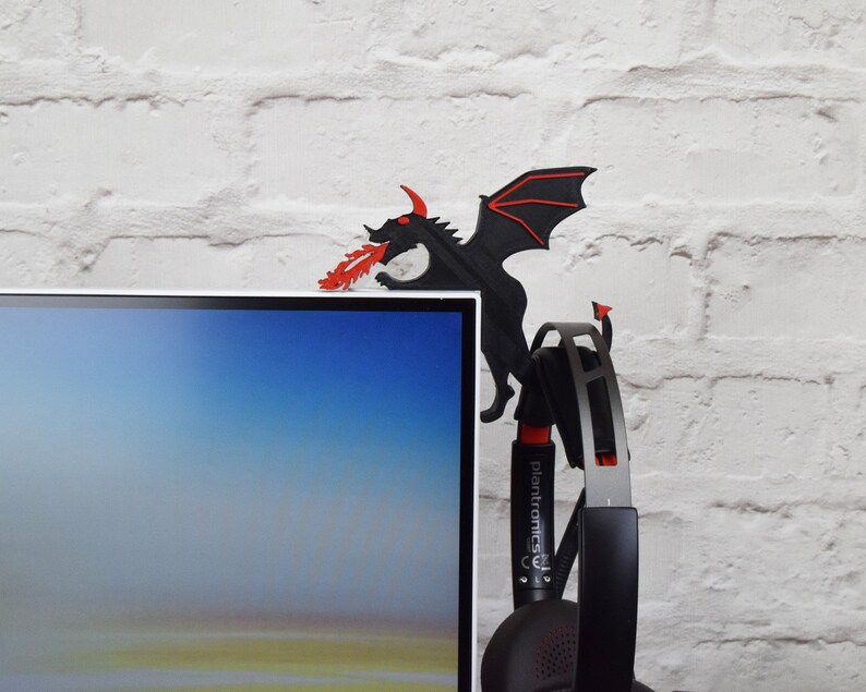 May include: A black dragon-shaped desk accessory with red accents, perched on the edge of a computer monitor. The dragon is spewing red flames. A black and silver headset hangs below the dragon.