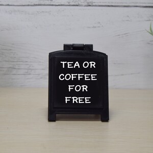 May include: A black A-frame chalkboard sign displaying the words "TEA OR COFFEE FOR FREE" in white. The sign is designed for tabletop use. The background features a light-coloured wooden surface, suggesting a cafe or shop setting.