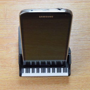 Puede incluir: Un tel&eacute;fono inteligente negro con la palabra "SAMSUNG" en la pantalla est&aacute; en un soporte de tel&eacute;fono negro con forma de piano. El soporte tiene teclas blancas.