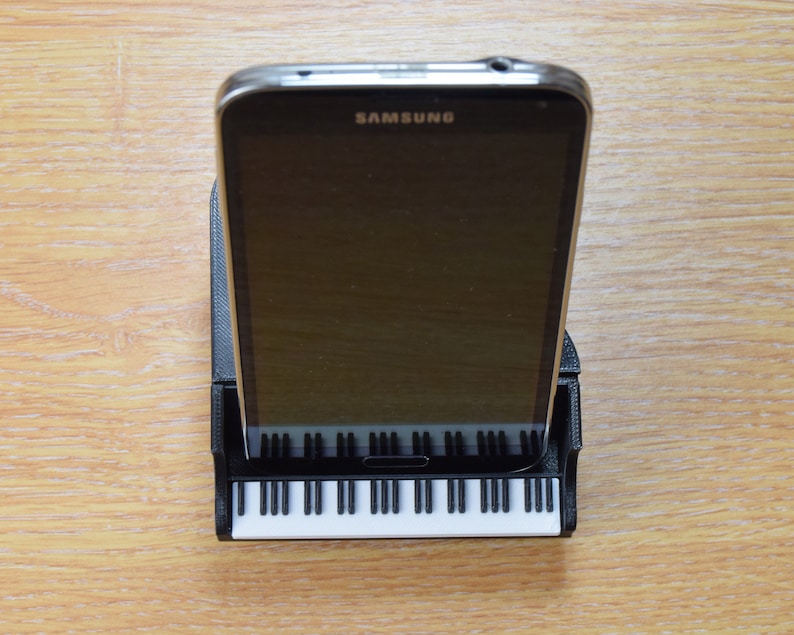 Puede incluir: Un tel&eacute;fono inteligente negro con la palabra "SAMSUNG" en la pantalla est&aacute; en un soporte de tel&eacute;fono negro con forma de piano. El soporte tiene teclas blancas.