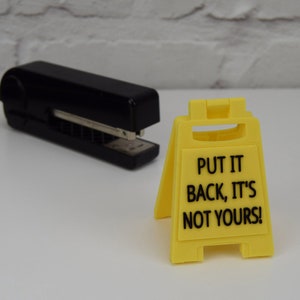 May include: A yellow plastic sign with the text "Put it back, it's not yours!" on a white surface. A black stapler is also in the image.