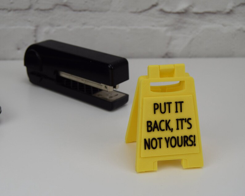 May include: A yellow plastic sign with the text "Put it back, it's not yours!" on a white surface. A black stapler is also in the image.