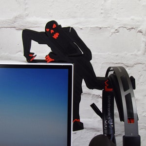 May include: A black 3D printed zombie figure with red eyes is perched on the top of a computer monitor. A pair of black headphones with a silver headband and red accents are hanging from the zombie's hand.
