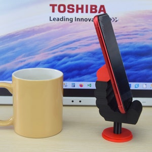 May include: A red smartphone is held upright in a black and red phone stand. The stand is shaped like a hand with a red base. The phone stand is on a wooden surface in front of a laptop with the Toshiba logo.