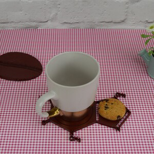 Puede incluir: Una taza de cer&aacute;mica blanca con un borde dorado se asienta sobre un posavasos hexagonal marr&oacute;n con una galleta de chocolate. El posavasos est&aacute; sobre un mantel a cuadros rojo y blanco.