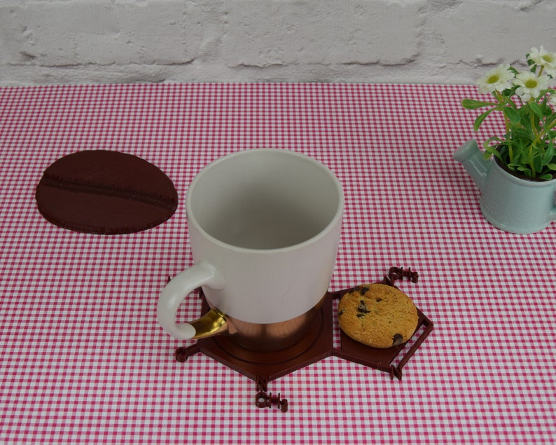 Puede incluir: Una taza de cer&aacute;mica blanca con un borde dorado se asienta sobre un posavasos hexagonal marr&oacute;n con una galleta de chocolate. El posavasos est&aacute; sobre un mantel a cuadros rojo y blanco.