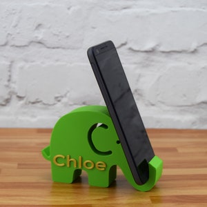May include: A green elephant phone stand with the name "Chloe" written on it. A black smartphone is propped up in the stand.