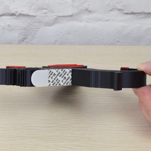 May include: Black plastic wall mount with a white adhesive strip. The adhesive strip has the text "WALL MOUNT" printed on it.