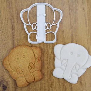 May include: Two white cookie cutters and a brown cookie in the shape of an elephant. The cookie cutters are detailed with the elephant's trunk, ears, and eyes.