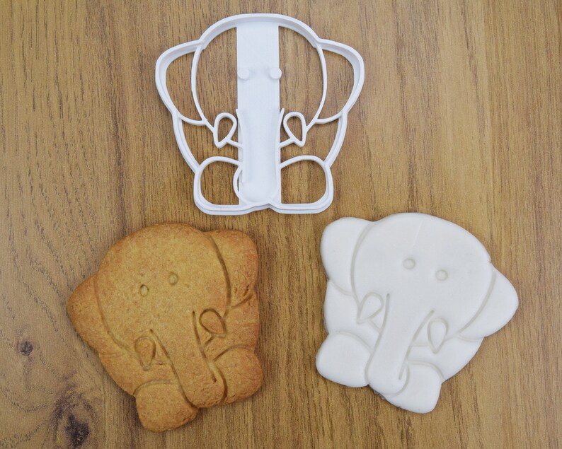 May include: Two white cookie cutters and a brown cookie in the shape of an elephant. The cookie cutters are detailed with the elephant's trunk, ears, and eyes.