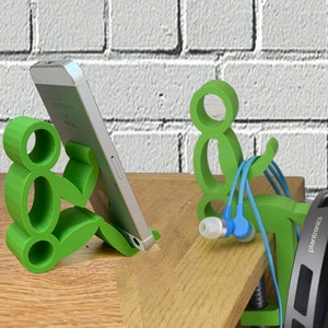 May include: Two green 3D printed phone and headphone holders. The phone holder is shaped like a person sitting with legs crossed. The headphone holder is shaped like a person sitting with legs straight out. The phone holder is holding a silver smartphone. The headphone holder is holding a pair of black headphones with the word "Plantronics" printed on the side.