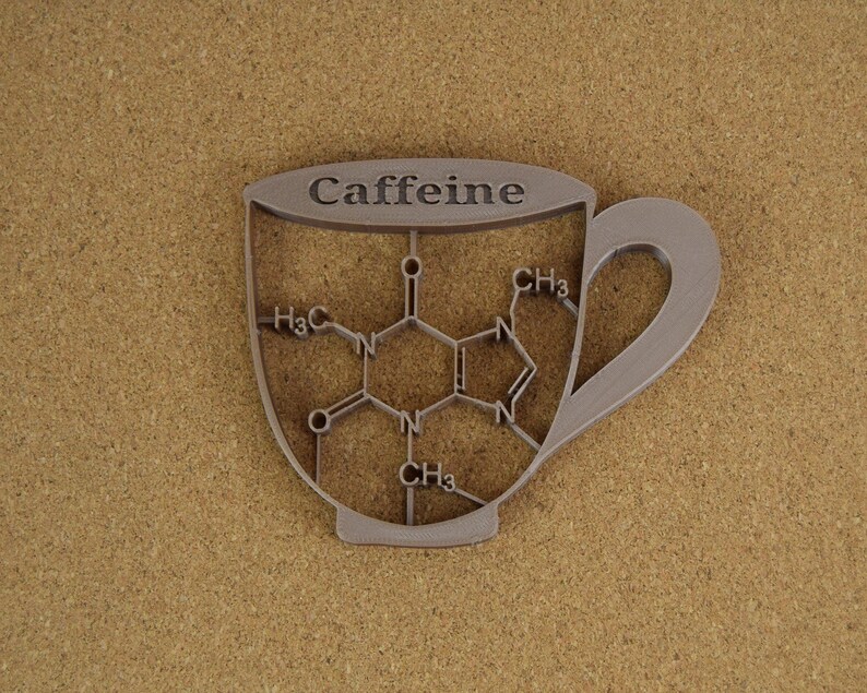 May include: A silver-colored, cup-shaped object with the word "Caffeine" at the top. The interior of the cup features a molecular structure diagram. The cup is set against a textured, tan background.