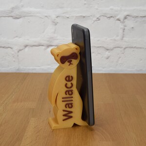 May include: A 3D printed phone stand shaped like a meerkat with the name "Wallace" printed on the side. The stand is holding a black smartphone.