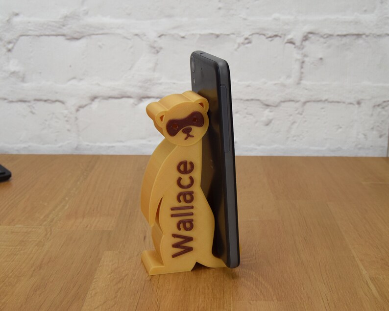May include: A 3D printed phone stand shaped like a meerkat with the name "Wallace" printed on the side. The stand is holding a black smartphone.