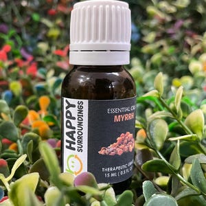 May include: A brown glass bottle of Myrrh essential oil with a white cap. The label reads "Happy Surroundings" and "Essential Oils Myrrh". The bottle contains 15 ml (0.5 fl oz) of therapeutic grade oil. The background is a mix of green and colorful foliage.