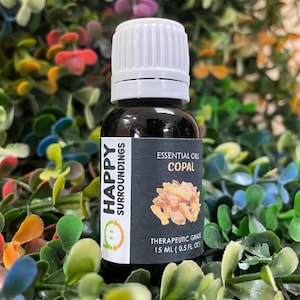 Puede incluir: Una botella de vidrio oscuro con tapa blanca, etiquetada "HAPPY SURROUNDINGS" y "ESSENTIAL OILS COPAL". La etiqueta también indica "THERAPEUTIC GRADE" y el volumen es de 15 ml.