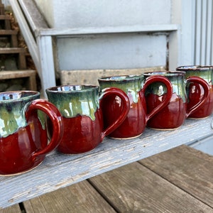 May include: A row of five handmade ceramic mugs. The mugs have a deep red base with a green and brown glaze at the top. Each mug has a large, curved handle. The mugs are displayed on a weathered wooden surface.