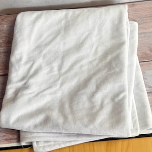 May include: A folded, white blanket made from a soft, textured material. The blanket is neatly folded and appears to be new. The blanket is a solid white colour.