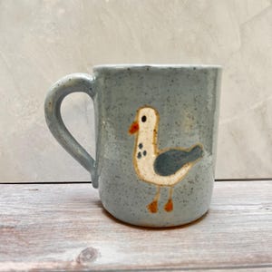 May include: A light blue ceramic mug with a speckled finish and a hand-painted illustration of a seagull on the front. The seagull has a white body, a dark gray wing, an orange beak, and orange feet, featuring a simple, rounded handle on the side.