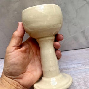 Puede incluir: Una copa de cerámica crema hecha a mano con un cuenco redondeado, un tallo largo y delgado y una base circular. La copa mide aproximadamente 15 cm de altura y probablemente se utiliza para beber bebidas.