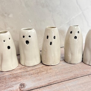 May include: Five off-white ceramic ghost-shaped vases arranged in a row. Each vase features a smooth, glossy finish with hand-painted black eyes and a circular mouth, creating a unique facial expression on every individual piece in the set.
