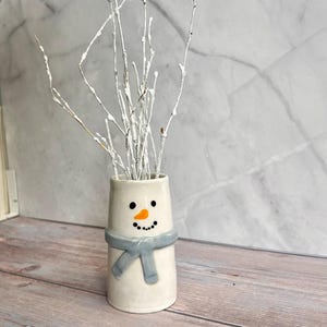 May include: A ceramic snowman vase with a light blue scarf and a smiling face, holding white-painted branches. The vase is on a wooden surface with a marble-patterned background. The snowman's face has black dots for eyes and a carrot nose.