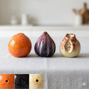 Op de afbeelding: Drie keramische fruitsculpturen: een oranje mandarijn, een vijg met paarse en zwarte tinten en een granaatappel met rode zaden. De sculpturen staan op een neutrale ondergrond en tonen hun gedetailleerde vakmanschap en levendige kleuren.
