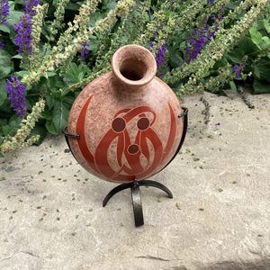 May include: A round, mottled tan ceramic vase with a narrow neck, featuring a stylized red graphic of three interconnected figures. The vase is supported by a black metal stand with three curved legs that cradle the base of the vessel.