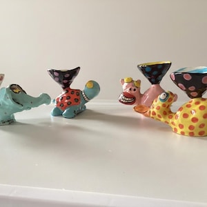 May include: Four ceramic egg cups shaped like whimsical animals including a light blue elephant, a blue and orange turtle, a pink pig, and a yellow duck. Each animal features a colourful, patterned bowl on its back with polka dot designs.