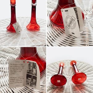 Könnte beinhalten: Rote Kerzenhalter und dekorative Flaschen aus Glas. Die Kerzenhalter haben einen ausgestellten Fuß und eine kleine Tasse für die Kerze. Die Flaschen haben einen bauchigen Boden und einen langen Hals. Jedes Stück hat ein Etikett mit der Aufschrift "AUTHENTIC Glassworks".